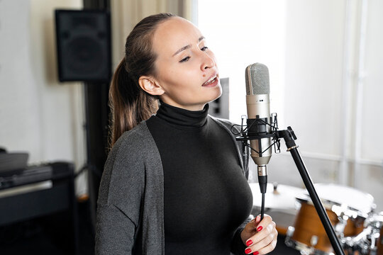 Young Asian or Caucasian brunette woman singing lyric song to microphone alone in professional music studio or concervatory class. Radio host speaking to her listeners in broadcasting radio studio. 