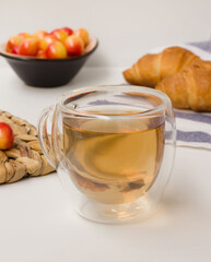 Morning herbal tea in a cup on a white table. Yellow-red cherries on a background in a bowl and two croissants on a towel