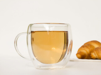 Glass cup with herbal tea on a white table. Two croissants in the background. Light breakfast on a summer morning