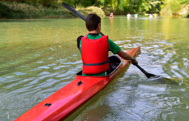 Un chico joven de espaladas en un primer plano haciendo piragüismo en el rio junto a vegetación frondosa. Actividades en la Naturaleza, desarrollo sostenible, turismo rural.