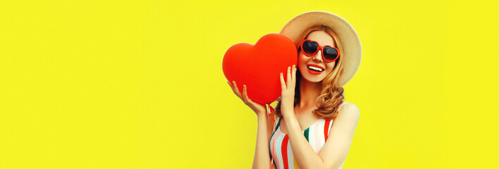 Portrait of happy smiling young woman with big red heart shaped balloon having fun wearing summer straw hat, sunglasses on yellow background, blank copy space for advertising text