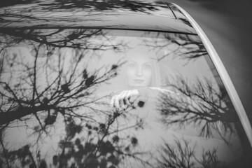 Art work. A black and white photo of a beautiful stylish girl sitting in a car. There are beautiful reflections on the windows of the car.