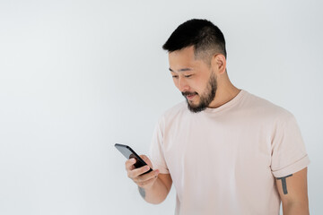 bearded asian man with tattoos using smartphone isolated on grey.