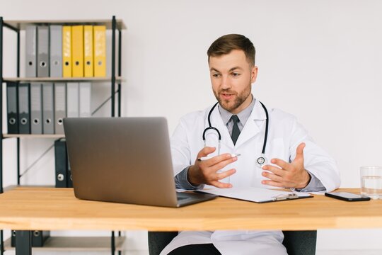 Happy Male Doctor Watching An Online Medical Webinar Or Training Seminar While Sitting With A Laptop In The Workplace.