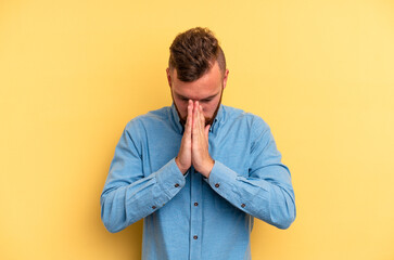 Young caucasian man isolated on yellow background praying, showing devotion, religious person looking for divine inspiration.
