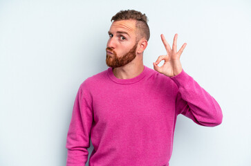 Young caucasian man isolated on blue background winks an eye and holds an okay gesture with hand.