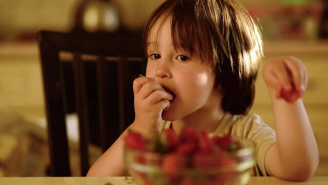 a three-year-old boy with long black hair is eating strawberries in the kitchen,