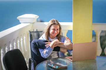 Young pretty business woman in blue shirt with laptop at sea view terrace is teaching online yoga