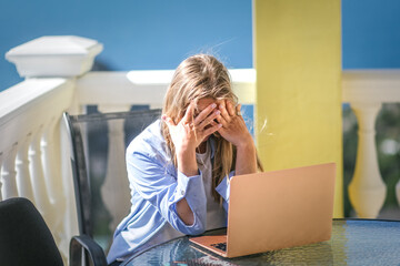 Upset tired freelance woman works on a sea view balcony with a laptop. She closes her face by hands