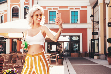 Young beautiful smiling hipster woman. Sexy carefree model posing on the street. Holding and drinking fresh vegetable cocktail smoothie drink in plastic cup with straw. Taking selfie photos