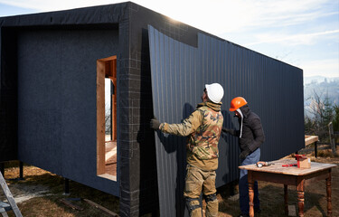 Male builders installing black corrugated iron sheet used as facade of future cottage. Men workers...
