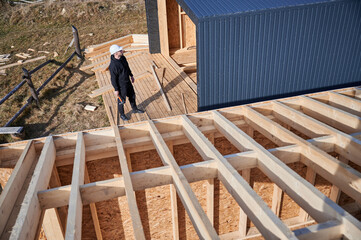 Male engineer building wooden frame house. Man developer on construction site, inspecting quality of work on sunny day, holding hammer in hands.