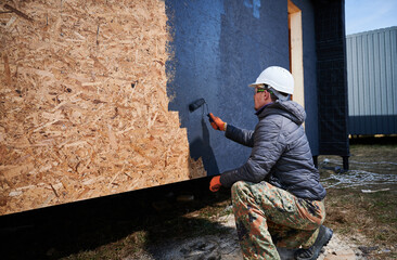 Male painter using paint roller, doing exterior paint work in a black color. Man worker building wooden frame house. Carpentry and construction concept.