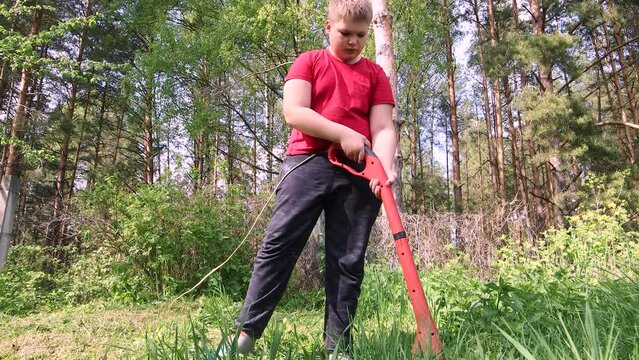 A boy mows tall green grass with an electric trimmer. The concept of caring for the territory of the house, garden plot, lawn. A man cuts weeds with a lawn mower. Warm soft light. UHD 4K.