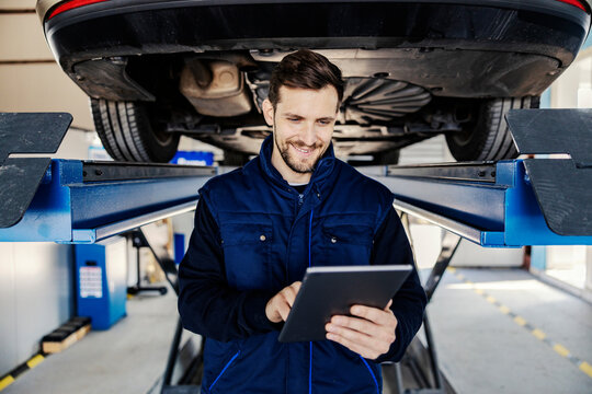 A smiling car service worker scrolling on tablet and checking on car in garage.