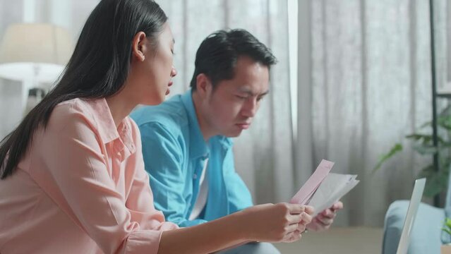 Close Up Of Asian Couple With A Laptop Looking At The Bill And Checking Money On The Purse Then Shaking Head And Having Headache
