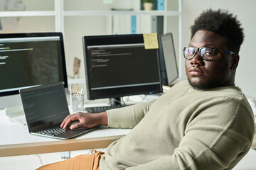 Serious African programmer looking at camera while sitting near computer monitor with script on screen