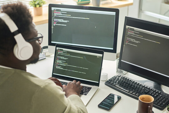 Rear view of African man in wireless headphones writing codes on computer sitting at workplace in IT office