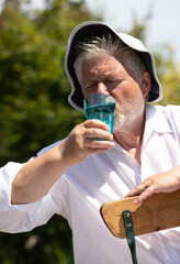 An elderly man in a sun hat sits in the garden in summer and sweats. He drinks water from a glass.
