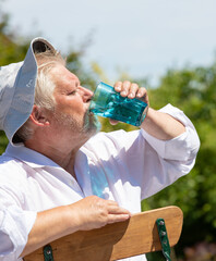 An elderly man in a sun hat sits in the garden in summer and sweats. He drinks water from a glass.