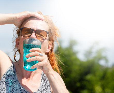 A woman suffers from high temperatures outdoors. She drinks mineral water from a glass.