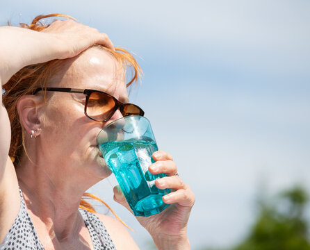 A woman suffers from high temperatures outdoors. She drinks mineral water from a glass.