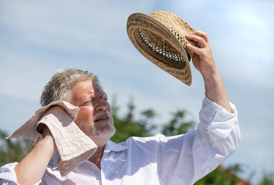 An elderly man sits outside in the sunshine. He is sweating and trying to protect himself from the sun's rays with a straw hat.