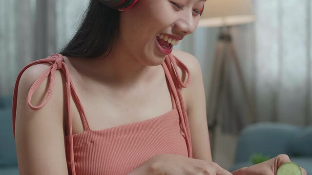 Close Up Of Smiling Asian Woman Listening To Music With Headphones And Singing While Slicing The Cucumber At Home
