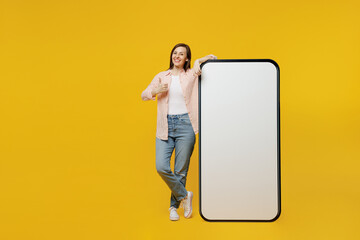 Full body young woman she 30s in striped shirt white t-shirt stand near big huge blank screen mobile cell phone with workspace copy space mockup area show thumb up isolated on plain yellow background.