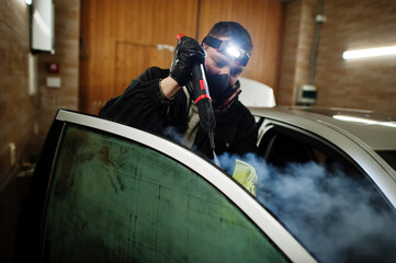 Man in uniform and respirator, worker of car wash center, cleaning car interior with hot steam cleaner. Car detailing concept.