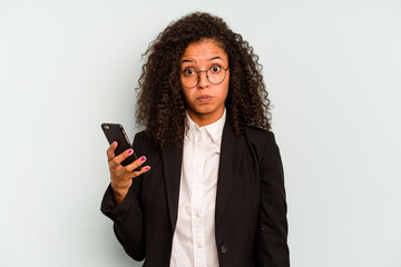 Young business Brazilian woman holding mobile phone isolated on white background shrugs shoulders and open eyes confused.