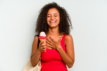 Young brazilian woman going to the beach holding an ice cream isolated on blue background laughs out loudly keeping hand on chest.