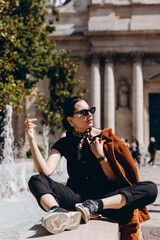 Portrait of a young stylish girl with black hair in glasses and a stylish brown jacket sitting near the fountain, a rebel.