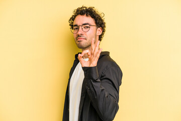 Young caucasian man isolated on yellow background winks an eye and holds an okay gesture with hand.