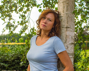 Thoughtful serious mature woman standing alone leaning against a birch tree.