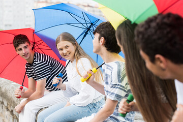 Blonde teenager girl talking with friends and holding colorful umbrellas in a rainy day.