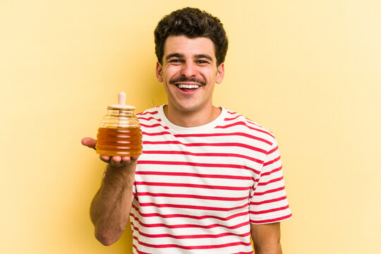 Young caucasian man holding honey jar isolated on yellow background laughing and having fun.
