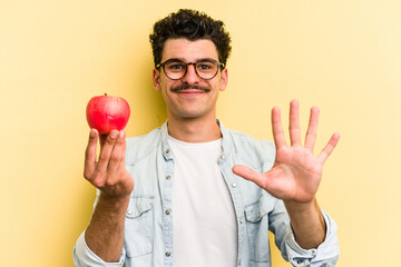 Young caucasian man holding an apple isolated on yellow background smiling cheerful showing number...