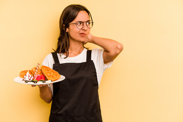 Young hispanic dependent woman holding waffles isolated on yellow background touching back of head, thinking and making a choice.