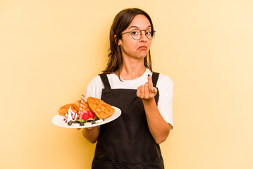 Young hispanic dependent woman holding waffles isolated on yellow background pointing with finger at you as if inviting come closer.