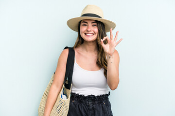 Young caucasian woman holding beach bag isolated on blue background cheerful and confident showing ok gesture.
