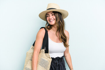 Young caucasian woman holding beach bag isolated on blue background looks aside smiling, cheerful and pleasant.