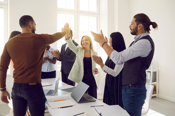 Mixed race team of business people celebrating result of successful teamwork. Happy young woman and man high five each other while coworkers are applauding, with sun lighting up room giving lens flare