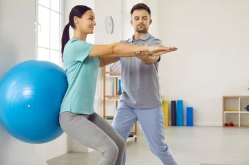 Foto op Plexiglas Rehabilitation therapy. Woman with back injury performs exercises with gymnastic ball during meeting with physiotherapist. Male doctor helps young woman who presses blue fitball with her back to wall. © Studio Romantic