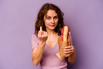 Young caucasian woman eating a sandwich isolated on purple background pointing with finger at you as if inviting come closer.