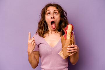 Young caucasian woman eating a sandwich isolated on purple background pointing upside with opened mouth.