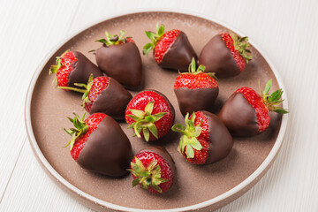 Ripe juicy strawberries dipped in dark chocolate on ceramic plate on light wooden table. Dessert background.