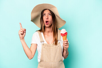 Young caucasian pregnant woman holding an ice cream isolated on blue background pointing upside with opened mouth.