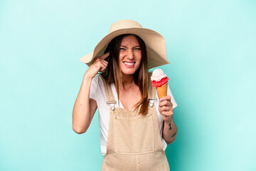 Young caucasian pregnant woman holding an ice cream isolated on blue background covering ears with...