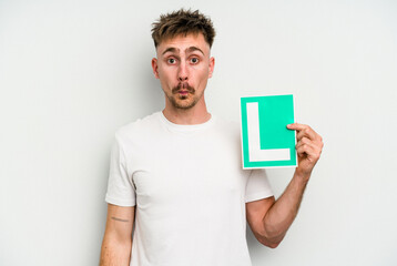 Young caucasian man holding L placard isolated on white background shrugs shoulders and open eyes...